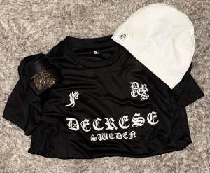 Decrese sweden  Outfit Paket – T-shirt + Bälte + Mössa - Decrese Outfit Paket – T-shirt + Bälte + Mössa Komplett streetwear-set som säljs tillsammans. T-shirt: svart med vit Decrese-text, sportig matchtröjekänsla, lätt och bekväm. Bälte: äkta läder, svart med silverfärgat spänne. Mössa: stilren svart med vit text. Perfekt paket för en modern och stilren look.