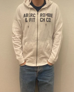Vit hoodie Abercrombie & Fitch M - Snygg vit hoodie från Abercrombie & Fitch i storlek M. Tröjan har dragkedja, snörning vid huvan och tryckt logga på bröstet. Tillverkad i mjuk bomull och har två fickor framtill. Perfekt för chill dagar och enkel att matcha med jeans.