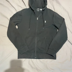 Svart hoodie från Polo Ralph Lauren - Svart hoodie från Polo Ralph Lauren med klassisk vit logga på bröstet. Tröjan har huva med snörning, dragkedja framtill och två fickor. Tillverkad i mjuk bomull, perfekt för chill dagar. Storlek S (passar XS–S). Skick 9/10 – inga fläckar eller hål. Nypris ca 1500–2000 kr. ✅ Äkta vara | 📩 Öppen för frågor | 🚚 Skickas inom 24 timmar 