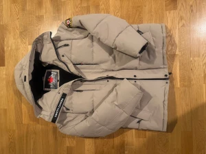 Beige pufferjacka från Zavetti Canada - Säljer en pufferjacka från Zavetti Canada i storlek L. Jackan har huva, svarta detaljer, patch på ärmen och broderad logga på insidan. Fylld med syntetmaterial och har dragkedja framtill. Perfekt för kalla dagar och har en clean, modern look. Köpt för 2500kr och säljer för 1800kr.(pris kan diskuteras)