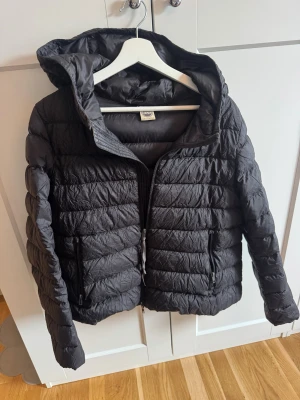 Svart pufferjacka från Colmar - Svart pufferjacka från Colmar med huva och dragkedja framtill. Jackan har quiltad design, två fickor med dragkedja och en lätt glansig yta. Perfekt för kyliga dagar och riktigt snygg till streetwear-stilen. Lite svagt utsvängd jacka.