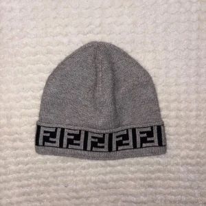 Grå Fendi mössa med logobård - Snygg grå mössa från Fendi med klassisk svart logobård runt kanten. Mössan är stickad och har en mjuk, skön känsla som passar perfekt när det är kallt ute. Fendi-loggan syns tydligt och ger en lyxig vibe till din look. Köpt på Vinted.