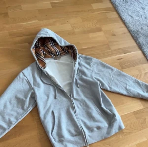 Grå Burberry hoodie med rutig huva - Säljer en grå hoodie från Burberry med dragkedja och snörning. Insidan av huvan har det klassiska Burberry-rutiga mönstret i beige, svart och rött. Tröjan har långa ärmar, två fickor fram och broderad logga på bröstet. Perfekt för dig som gillar stilrena streetwear-plagg.