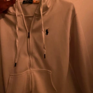 Vit zip up hoodie från Polo Ralph Lauren - Säljer en vit hoodie från Polo Ralph Lauren med dragkedja och snörning i huvan. Tröjan har lång ärm, ficka framtill och den klassiska Polo-loggan broderad på bröstet. Perfekt för chill dagar och enkel att matcha med jeans eller joggers.