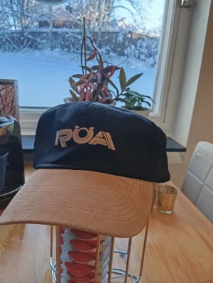 Svart och beige keps från ROA - Snygg keps från ROA med svart krona och beige skärm. Framtill finns en vit ROA-logga i coolt tryck. Kepsen har klassisk form och passar perfekt för dig som vill ha en stilren accessoar med streetkänsla. Skärmen är böjd och materialet känns slitstarkt.