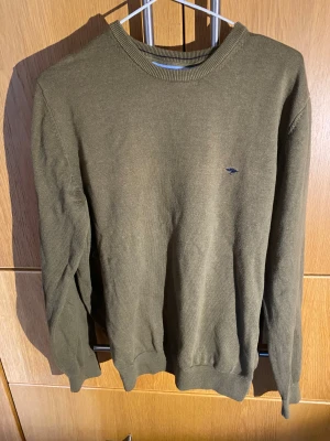Olivgrön stickad tröja Fynch-Hatton M - Olivgrön stickad tröja från Fynch-Hatton i mjuk bomull. Klassisk rund hals och ribbade muddar vid ärmslut och nederkant. Liten broderad logga i mörkblått på bröstet. Perfekt för dig som gillar stilren och enkel design.