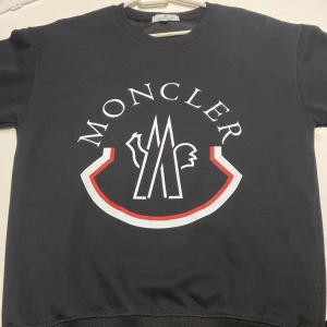 Svart sweatshirt från Moncler med tryck - Svart sweatshirt från Moncler i 100% bomull med stort vitt och rött logotryck på bröstet och klassisk Moncler-patch på ärmen. Rund hals och ribbade muddar ger en clean och stilren look. Perfekt för dig som gillar exklusiva streetwear-vibes. Den passar för både L och M