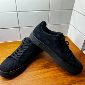 Svarta mocka sneakers med platt sula - Snygga helsvarta sneakers från Selected homme med ovandel i mjuk mocka och platt sula. Skorna har rund tå, svarta snören och en stilren, minimalistisk look som passar till det mesta. Perfekt för dig som gillar enkel och clean stil. Priset är ristat i sten går ej att pruta!!