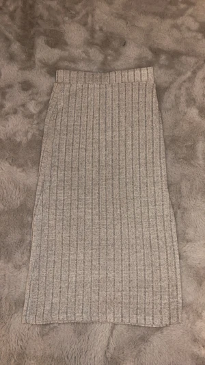 Grå ribbad långkjol - Säljer en grå långkjol med ribbad struktur och vertikala ränder. Kjolen har en rak och enkel form som ger en clean look. Perfekt att matcha med sneakers eller boots för en chill vibe.