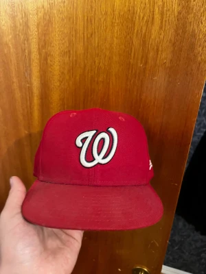 Röd New Era Washington Nationals keps - Säljer en röd keps från New Era med Washington Nationals-logga broderad framtill i vitt och svart. Klassisk basebollmodell med böjd skärm och MLB-logga baktill. Kepsen är tillverkad i bomull och har en stilren look som passar perfekt till streetwear.