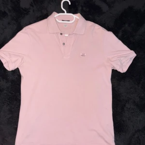  pikétröja C.P. Company - Snygg ljusrosa pikétröja från C.P. Company med klassisk krage och två svarta knappar framtill. Tröjan har diskret logga på bröstet och är gjord i mjuk bomull som känns skön mot huden. Perfekt för dig som gillar  stilrena och enkla plagg. Självklart äkta