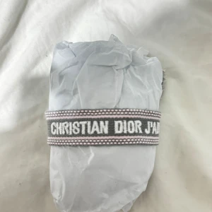 Christian Dior J'Adior armband - Snyggt armband från Christian Dior J'Adior i grått med ljusrosa detaljer och vit broderad text. Armbandet är gjort i textil och har två flätade snören med tofsar för justerbar passform. Perfekt statement piece med lyxig vibe.