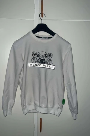 Vit sweatshirt från Kenzo med tiger - Säljer en vit sweatshirt från Kenzo i storlek M med det ikoniska tigermotivet och 'Kenzo Paris' broderat på bröstet. Tröjan har ribbade muddar vid hals, ärmslut och nederkant samt en grön Kenzo-tag på sidan. Perfekt för dig som gillar streetwear och snygga loggor.
