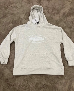 Corteiz hoodie med tryck i storlek L - Snygg ljusgrå hoodie från La Bett med ett vitt tryck framtill som föreställer en byggnad och stjärnor. Tröjan har huva, ribbade muddar och en klassisk magficka. Perfekt för dig som gillar enkel och stilren streetwear.