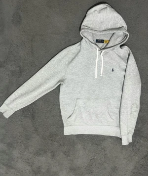 Grå hoodie från Polo Ralph Lauren - Snygg ljusgrå hoodie från Polo Ralph Lauren med klassisk broderad logga på bröstet. Tröjan har huva med vit snörning, magficka och ribbade muddar. Perfekt för chill dagar och enkel att matcha med jeans eller joggers.