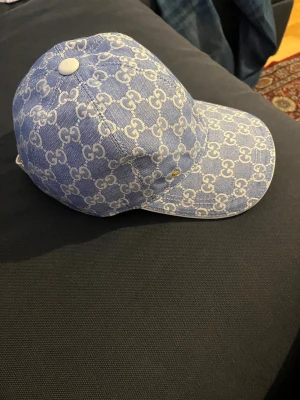 Blå Gucci keps med GG-mönster - Säljer en blå keps från Gucci med ikoniskt vitt GG-mönster över hela kepsen. Kepsen har en böjd skärm, vit knapp på toppen och justerbart spänne bak i vitt. Materialet är bomull och kepsen har en liten guldfärgad GG-detalj framtill. Perfekt för dig som vill sticka ut med lyxig streetstyle.