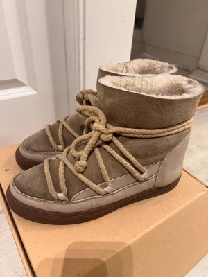 Beige INUIKII boots med snörning - Säljer ett par snygga beige boots från INUIKII med grov snörning och platt brun sula. Utsidan är i mocka och skinn, insidan är fodrad med mjuk päls för extra värme. Perfekta för kalla dagar och har en unik design med rund tå.