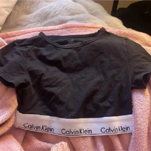 Svart croppad topp Calvin Klein - Svart croppad topp från Calvin Klein med korta ärmar och vit elastisk resår nertill med logga runt om. Perfekt till jeans eller shorts för en sportig och avslappnad look.