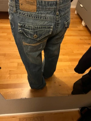 Blå wide jeans från Blueoutset - Säljer ett par blå jeans från Blueoutset med bred passform och raka ben. Jeansen har coola detaljer som stora bakfickor med knappstängning och slitningar. Materialet är klassisk denim och midjan är normal. Perfekt för dig som gillar avslappnad stil.