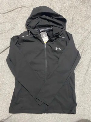 Helt ny Svart hoodie från Under Armour M - Svart hoodie från Under Armour med silverfärgat logotryck på bröstet och stor UNDER ARMOUR-text på ryggen. Tröjan har dragkedja framtill, huva och är tillverkad i lätt polyester med HeatGear-teknologi som håller dig sval och torr. Perfekt för träning och aktiva dagar. Helt äkta från Under armour 