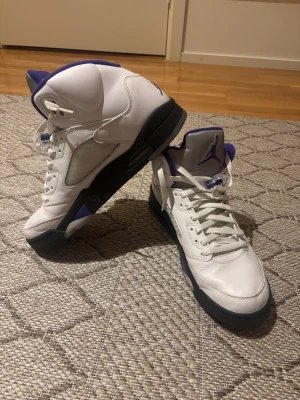 Nike Air Jordan 5 Retro vit/lila - Nike Air Jordan 5 Retro sneakers i vit skinn med lila detaljer och svart sula. Klassisk Jumpman-logga på tungan och hälen, samt genomskinlig sula med Jordan-tryck och högt skaft. Perfekt för dig som vill sticka ut med en ikonisk basketstil.