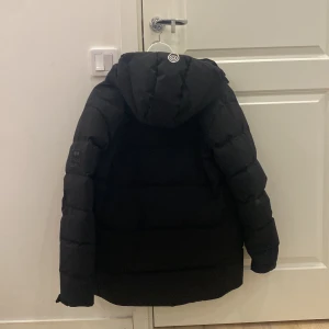 Svart pufferjacka från Sail Racing - Svart pufferjacka från Sail Racing med huva och två bröstfickor med dragkedja. Jackan har en stilren look med Sail Racing-logga på bröstet och patch på ärmen. Perfekt för kalla dagar och har en skön, vadderad känsla. Bästa vinterjackan man kan ha under de kalla vintertiderna, denna jackan är perfekt för dig som söker något varmt, stilren och perfekt till vintern för ett lågt pris på 1000kr. Jackan har använts ett par gånger och är i perfekt skick utan skador eller besvär, möts upp i Stockholm