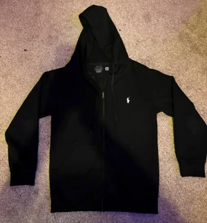 Svart hoodie från Polo Ralph Lauren - Svart hoodie från Polo Ralph Lauren med vit broderad logga på bröstet. Tröjan har huva med snörning, dragkedja framtill och två fickor. Slim fit-modell i mjuk bomull som är perfekt för chill dagar. Klassisk och stilren design som alltid funkar.