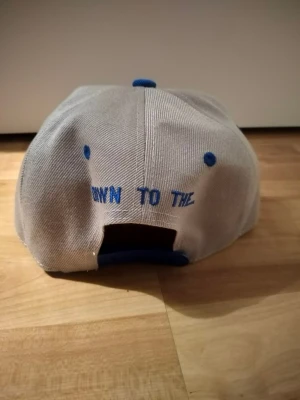 Grå och blå Brooklyn keps Soxxx - Snygg keps i grått med blå detaljer och broderad text 'BROOKLYN' framtill. Platt blå skärm, blå ventilationshål och knapp på toppen. Justerbar baktill med blå snapback. Broderad basebollhandske framtill. Perfekt för dig som gillar streetstyle.