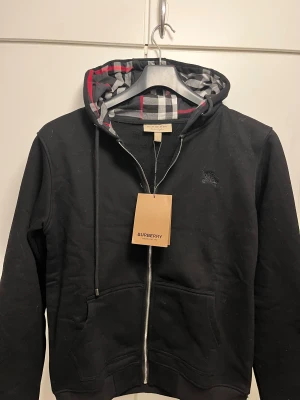 Svart Burberry hoodie med dragkedja - Svart hoodie från Burberry med klassiskt rutigt foder i huvan och snyggt broderat Burberry-emblem på bröstet. Tröjan har dragkedja framtill, snörning vid huvan och två fickor. Materialet är mjuk bomull och polyester, perfekt för dig som gillar stilren streetwear.