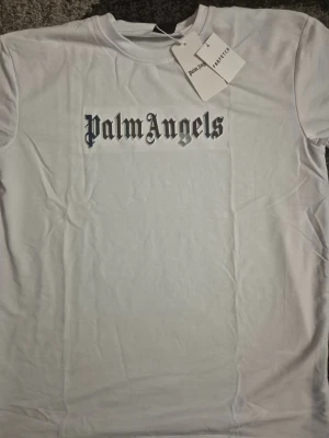Vit Palm Angels t-shirt XL - Vit t-shirt från Palm Angels i storlek XL med klassisk rund hals och tryckt logga i svart och silver på bröstet. T-shirten är gjord i mjuk bomull och har en clean, stilren look som passar perfekt till streetwear. Snyggt tryck med gotisk font framtill. Helt ny och perfekt