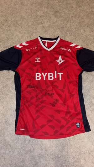 Astralis officiell CS tröja Hummel XL - Röd och mörkblå Astralis Officell CS tröja från Hummel i funktionsmaterial. Signerad av BLAMEF,STAEHR,BUZZ, med flera röjan har korta ärmar, vit V-ringad krage och tryckta loggor samt autografer på framsidan. Snyggt geometriskt mönster på bröstet och klassiska Hummel-pilar på axlarna.
