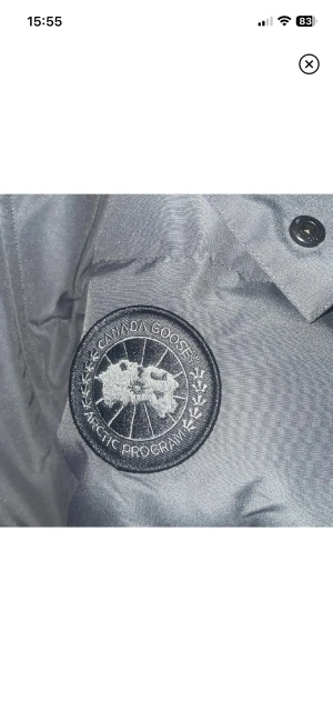 Mörkgrå pufferjacka från Canada Goose - Säljer en mörkgrå pufferjacka från Canada Goose med snyggt broderat logomärke på ärmen. Jackan har huva, dragkedja och knappar framtill samt två praktiska sidofickor. Perfekt för kalla dagar och har en clean, modern look.