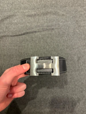 Svart  skärp med H-spänne - Snyggt svart skärp med ett stort, silvrigt H-spänne i metall. Bältet är gjort i äkta skinn med vit kontrastsöm längs kanterna. Perfekt accessoar för att lyfta din outfit med en lyxig touch och klassisk design.