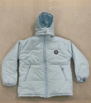 Ljusblå pufferjacka från Trapstar - Säljer en ljusblå pufferjacka från Trapstar med huva och dragkedja framtill. Jackan har två sidofickor med dragkedja, elastiska ärmslut och stor Trapstar-logga i svart och vitt på ryggen. Perfekt för kalla dagar och har en clean streetwear-vibe.