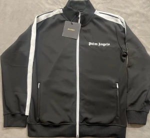 Svart tröja Palm Angels - Palm Angels svart trackjacka med vita ränder längs ärmarna och vit dragkedja framtill. Jackan har ribbade muddar, ståkrage och logotypen broderad på bröstet. Snygg och sportig design som sticker ut med klassiska detaljer.