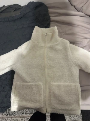Vit teddyjacka från 157, storlek S - Mysig vit teddyjacka från 157 med hög krage och två stora fickor framtill. Jackan har hel dragkedja och är tillverkad i fluffigt fleece-material som ger en chill vibe. Perfekt för kalla dagar när du vill vara både varm och snygg.