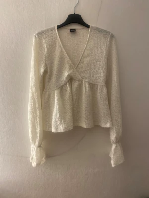 Vit omlottblus med volang från Gina Tricot - Supersöt vit blus från Gina Tricot i storlek XS. Blusen har omlott-design med v-ringning, volang nedtill och långa ärmar med knyt vid ärmslut. Materialet är strukturerat och känns mjukt mot huden. Perfekt för dig som gillar romantiska detaljer och en feminin look.