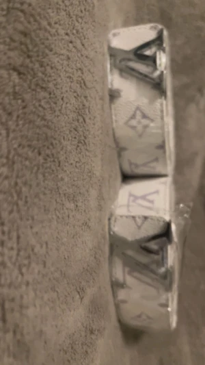 Vit bälte med monogram - Säljer ett snyggt vitt bälte  med klassiskt monogrammönster i silver och ljuslila. Bältet har en stilren, fyrkantig form och en stor spännet. Perfekt för dig som vill ha en exklusiv accessoar med lyxig känsla.