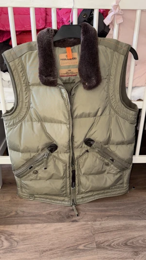 Olivgrön dunväst från Parajumpers - Snygg olivgrön dunväst från Parajumpers med fluffig mörkbrun krage i pälsimitation. Västen har flera dragkedjeförsedda fickor, coola detaljer och klassisk logga bak i nacken. Perfekt för lager-på-lager under kyliga dagar. Kemtvätad och knappt använd!