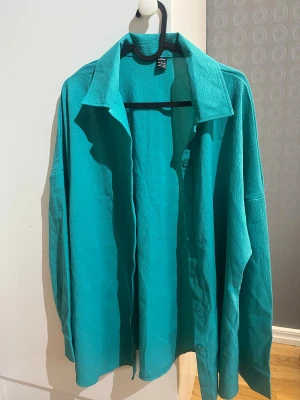 Turkos skjorta från SHEIN - Turkos skjorta från SHEIN med krage och knappar framtill. Passar 40 /42. Skjortan har en loose passform, långa ärmar och är tillverkad i ett lätt, strukturerat syntetmaterial. Perfekt för dig som gillar färg och vill ha en chill vibe i garderoben. 