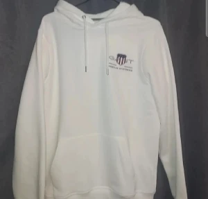Vit hoodie från GANT - Vit hoodie från GANT med broderad logga och sköld på bröstet. Tröjan har huva med snörning, stor magficka och långa ärmar. Mjuk och skön bomullskvalitet, perfekt för chill dagar. Snygga detaljer på snörningen med metalländar.
