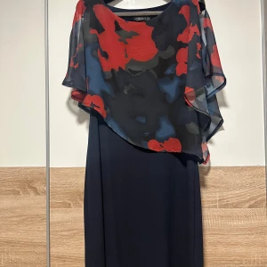 Marinblå långklänning med blommigt overlay - Snygg marinblå långklänning från Wear house med ett transparent overlay i blommigt mönster i rött, blått och svart. Klänningen har en loose passform och är ärmlös med ett elegant fall. Perfekt för dig som vill sticka ut med färg och stil.