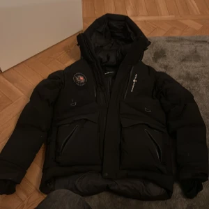 Svart dunjacka Sail Racing Antarctica - Säljer en svart Sail Racing Antarctica Expedition dunjacka med huva och flera praktiska fickor med dragkedja. Jackan har Sail Racing-logga på bröstet och patch på axeln. Materialet är polyester och polyamid med dunfyllning, perfekt för kalla vinterdagar.