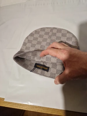 Louis Vuitton rutig mössa i grått - Snygg mössa från Louis Vuitton i grå och beige ton med klassiskt rutigt mönster. Mössan har en bred uppvikt kant med LV-logga framtill och är tillverkad i mjukt stickat material. Perfekt accessoar för dig som gillar exklusiv streetstyle.