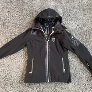 Svart softshelljacka med huva från J.Lindeberg - Svart softshelljacka från J.Lindeberg med huva och dragkedja framtill. Jackan har två framfickor med dragkedja, logotyp på bröstet och detaljer i blankt material. Insidan har kontrasterande foder med J.Lindeberg-tryck.