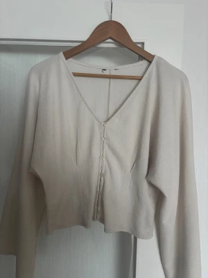 Beige blus med v-ringning och knappar - Säljer en stilren beige blus med v-ringad hals och knappar framtill. Blusen har lång ärm och är croppad i modellen, vilket ger en modern och avslappnad vibe. Materialet är lätt och har en diskret struktur som gör den extra snygg. Lappen är bortklippt men det är storlek M