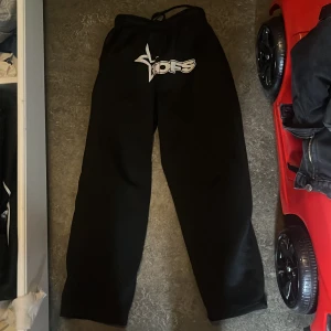 Svarta mjukisbyxor från Nofs - Svarta mjukisbyxor från Nofs med vit logga och stjärndetalj framtill. Byxorna har snörning i midjan och en loose passform med raka ben. Perfekta för chill eller träning, tillverkade i mjukt material för maximal komfort.