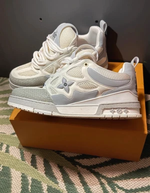 Louis Vuitton vita sneakers med mesh - Säljer ett par fräscha Louis Vuitton sneakers i vitt och ljusgrått. Skorna har meshpaneler, skinn och syntetdetaljer, samt LV-blomman på sidan och logga på plösen. Rund tå, platt sula och snörning. Kommer med originalkartong.