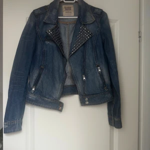 Blå jeansjacka med nitar från Zara - Snygg blå jeansjacka från Zara Trafaluc med coola svarta nitar på kragen och två dragkedjefickor framtill. Jackan har klassisk krage, knappdetaljer och är långärmad. Perfekt för dig som vill ha en edgy stil.