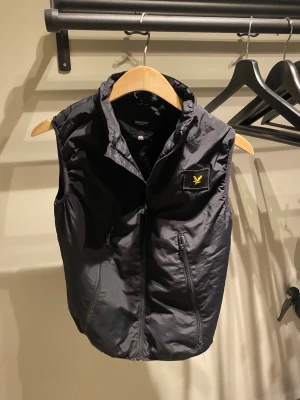 Svart väst Lyle & Scott Junior - Svart dunväst från Lyle & Scott Junior med gul logotyp på bröstet. Västen har två praktiska fickor med dragkedja och hög krage. Designen är stilren och sportig, perfekt för att addera lite edge till din outfit. 